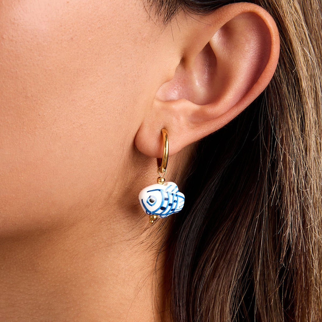 Close-up of an ear wearing a gold hoop earring with a blue and white fish charm.