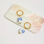 Pair of earrings with blue floral design on white beads, displayed on a shell.
