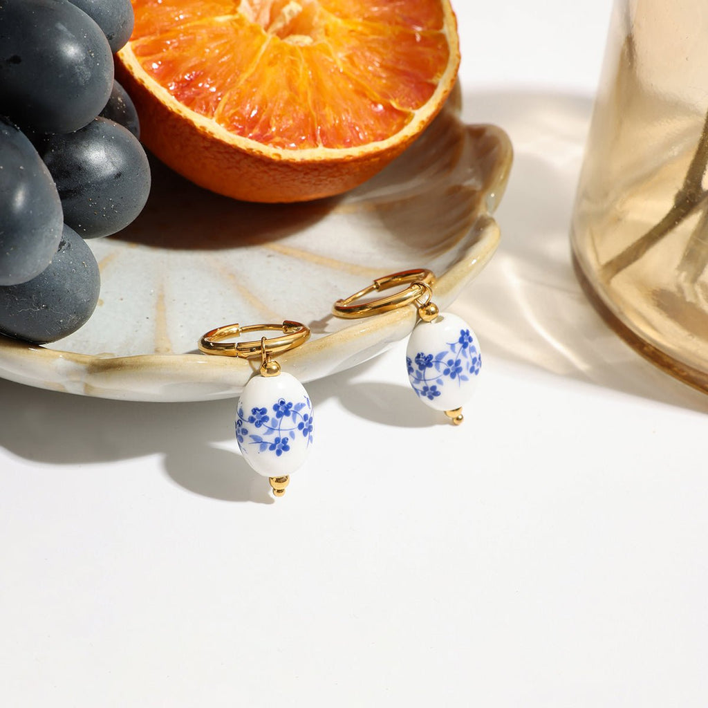 Pair of earrings with floral designs on a white surface with grapes and an orange in the background