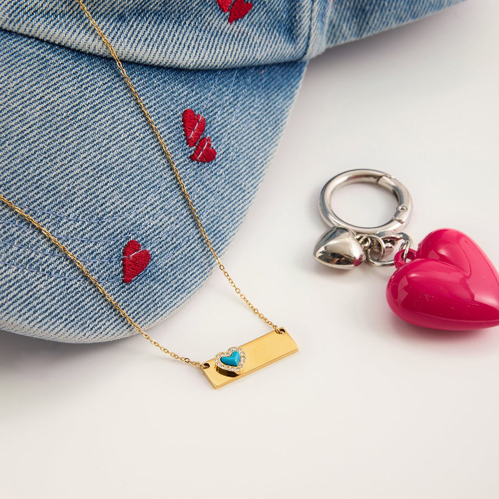 Gold necklace with heart pendant, denim jacket with red hearts, and pink heart keychain on a white surface