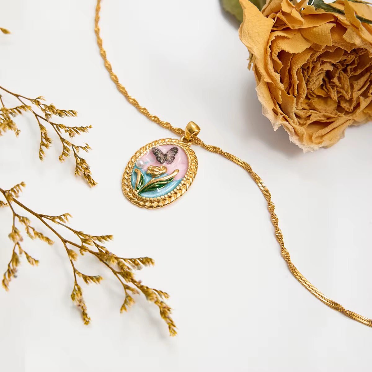 Gold necklace with a pendant featuring a butterfly and flower design, placed on a white surface with dried flowers.