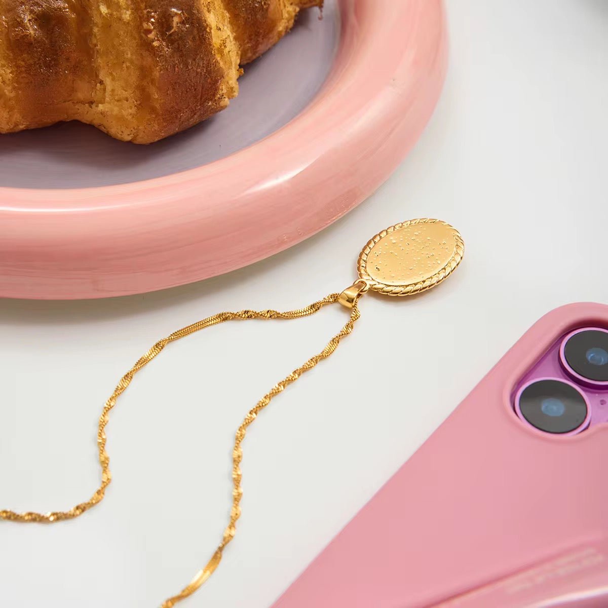 Gold phone chain on a pink phone with a croissant on a pink plate in the background