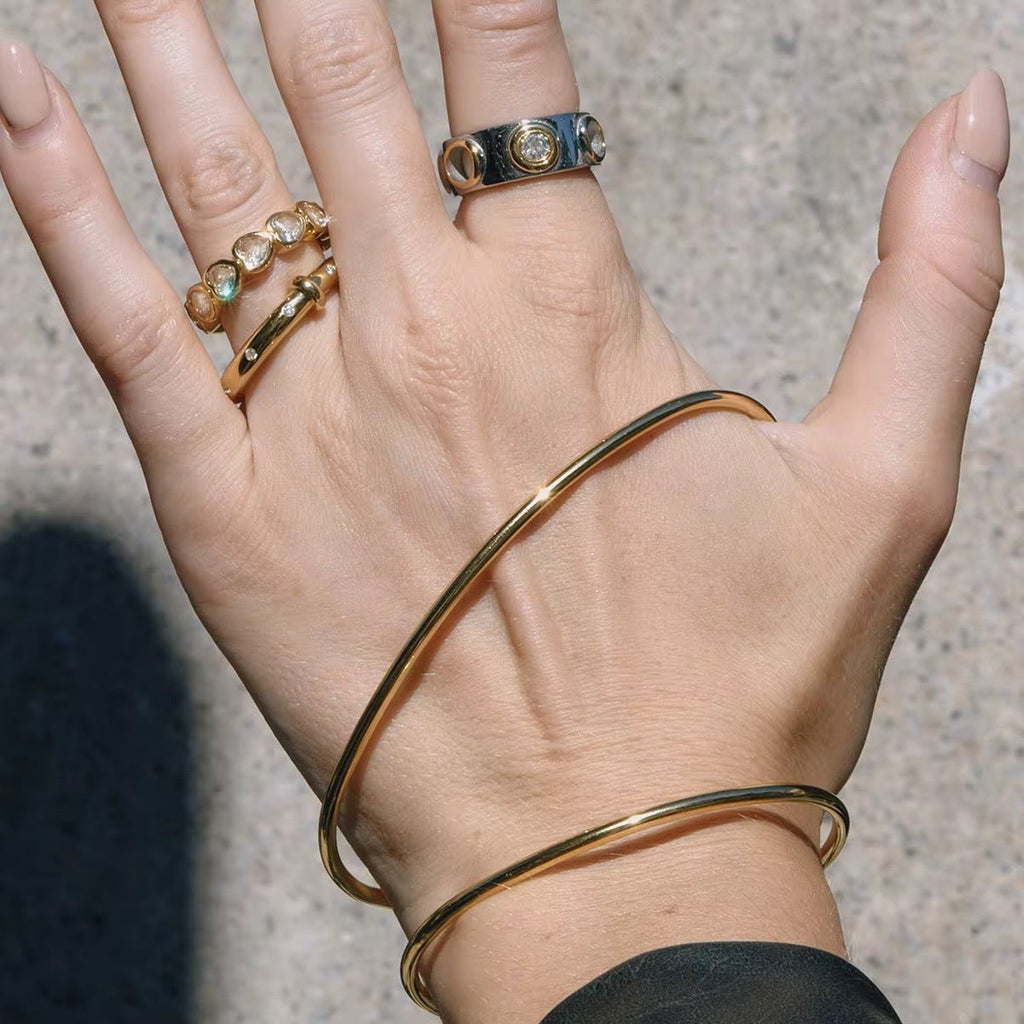 Hand wearing multiple gold bracelets and rings on a neutral background