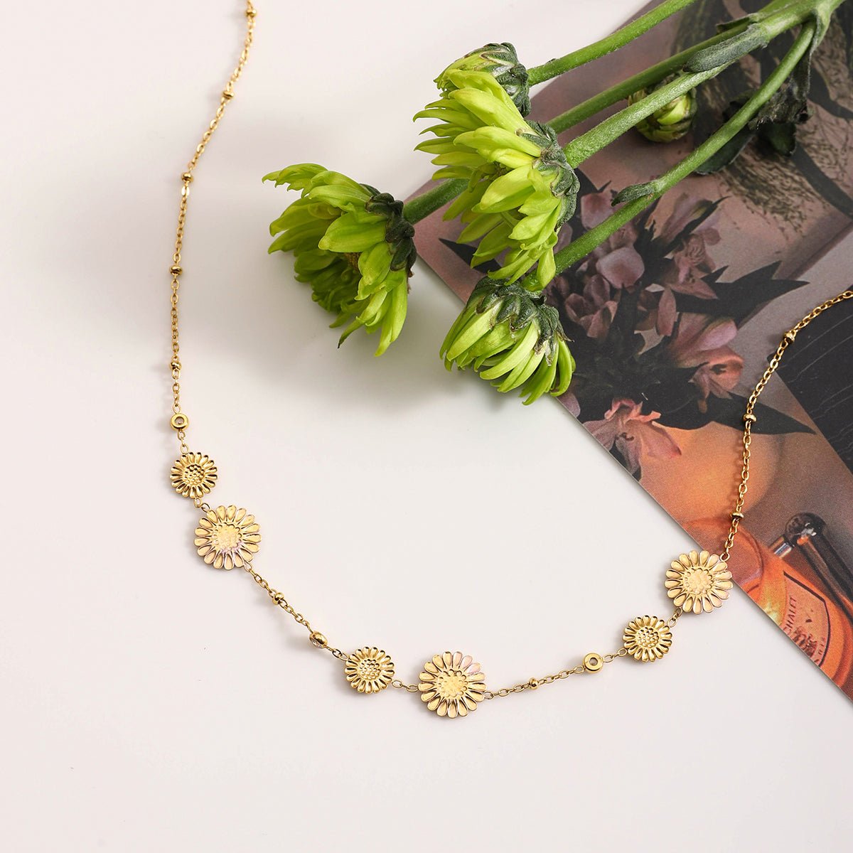 Gold necklace with sunflower designs on a white surface with green flowers and a magazine.