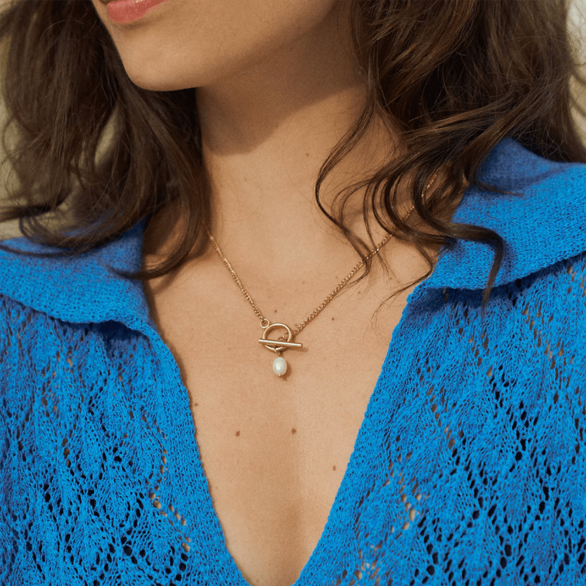 Close-up of a person wearing a gold necklace with a pearl pendant, against a blue textured background.