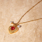 Gold necklace with a heart-shaped pendant and red stone on a beige background