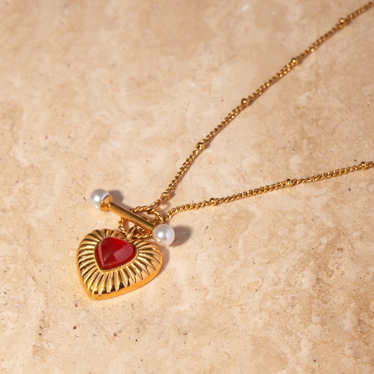Gold necklace with a heart-shaped pendant and red stone on a beige background