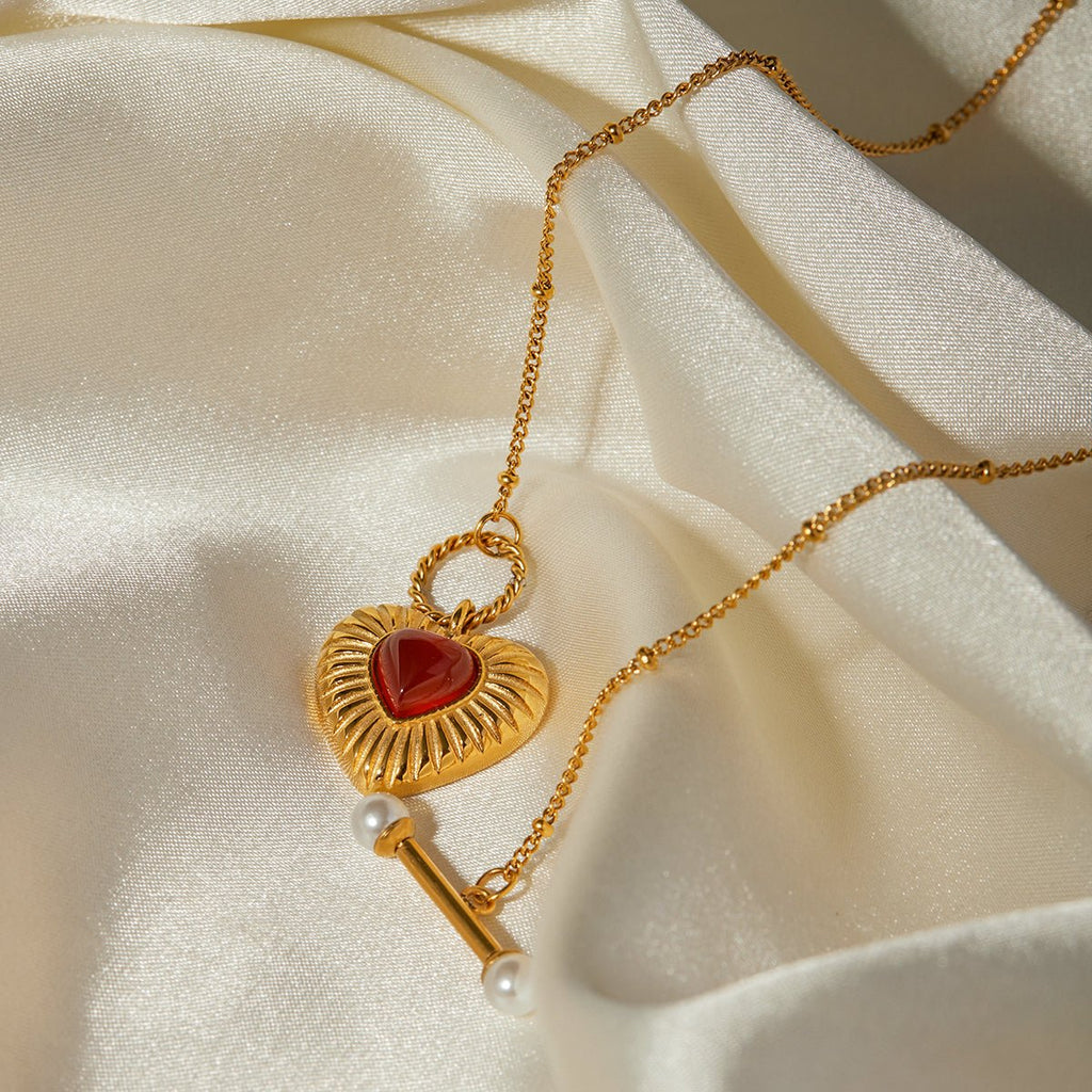 Gold necklace with a heart-shaped pendant and red stone on a beige fabric background