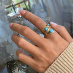 Hand wearing a gold ring with turquoise stones against a blurred indoor background