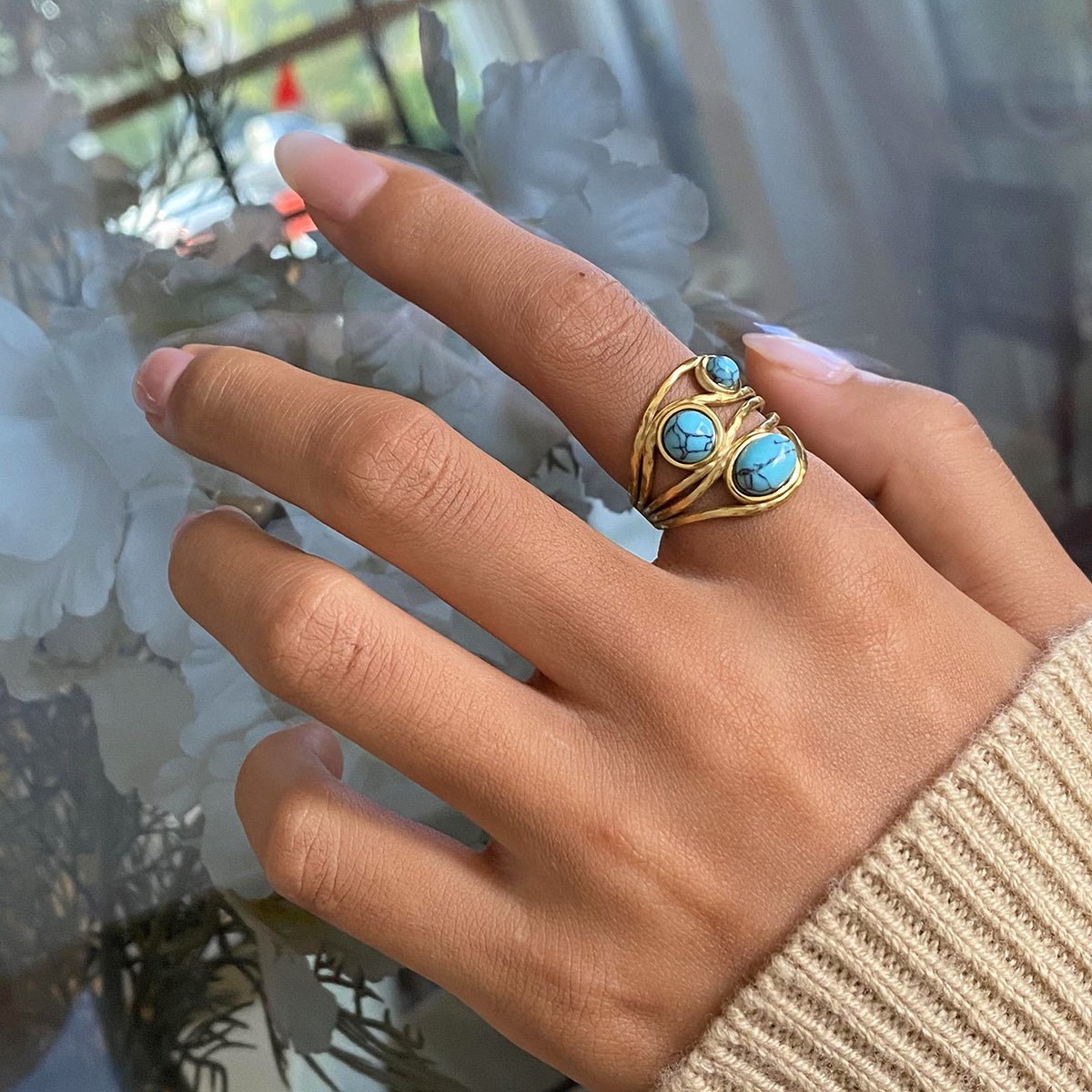 Hand wearing a gold ring with turquoise stones against a blurred indoor background