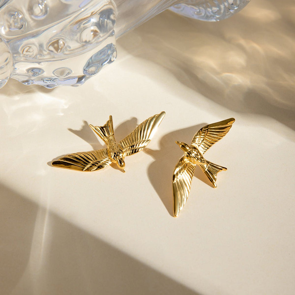 Gold bird-shaped earrings on a beige surface with a crystal object in the background