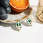 Pair of earrings with green and white design on a marble surface with fruit and glassware.
