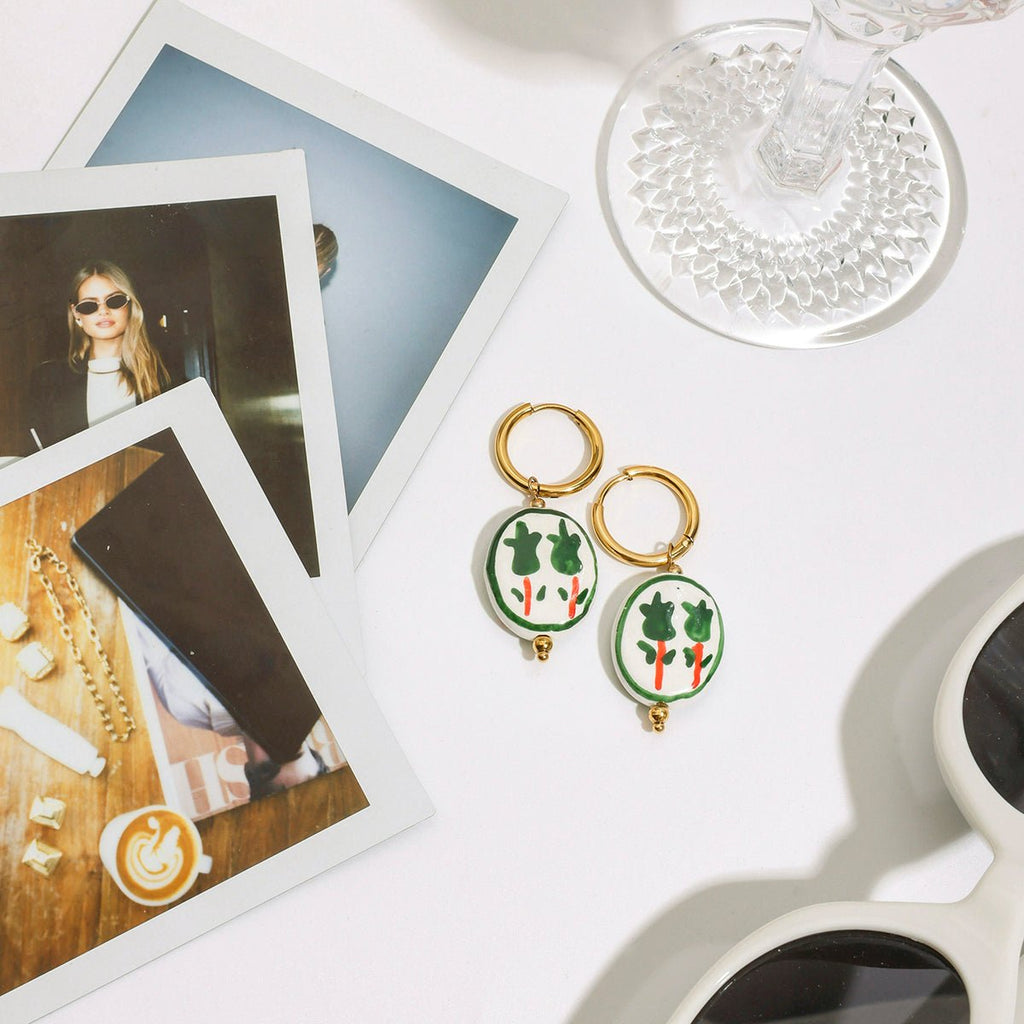 Pair of earrings with green and gold design on a white surface with photos and sunglasses.