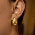 Close-up of an ear wearing a gold earring with a clear gemstone on a neutral background
