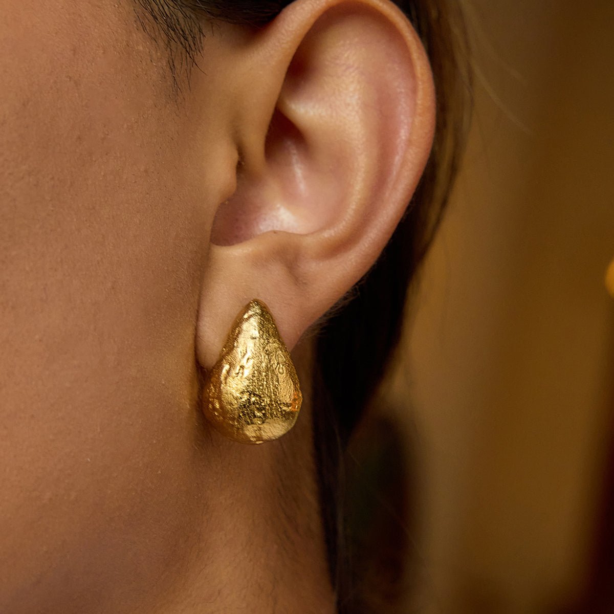 Close-up of an ear wearing a gold earring with a clear gemstone on a neutral background