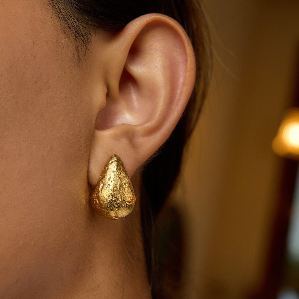 Gold teardrop earring worn on an ear with a blurred background