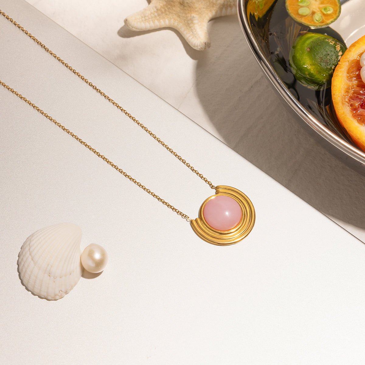 Gold necklace with a pink pendant on a white surface with a shell and fruit in the background
