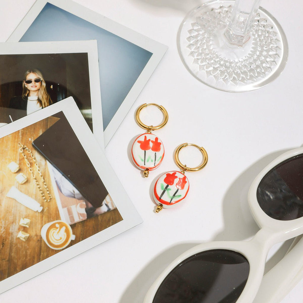 Pair of earrings with red floral design on a white surface with photos and sunglasses.