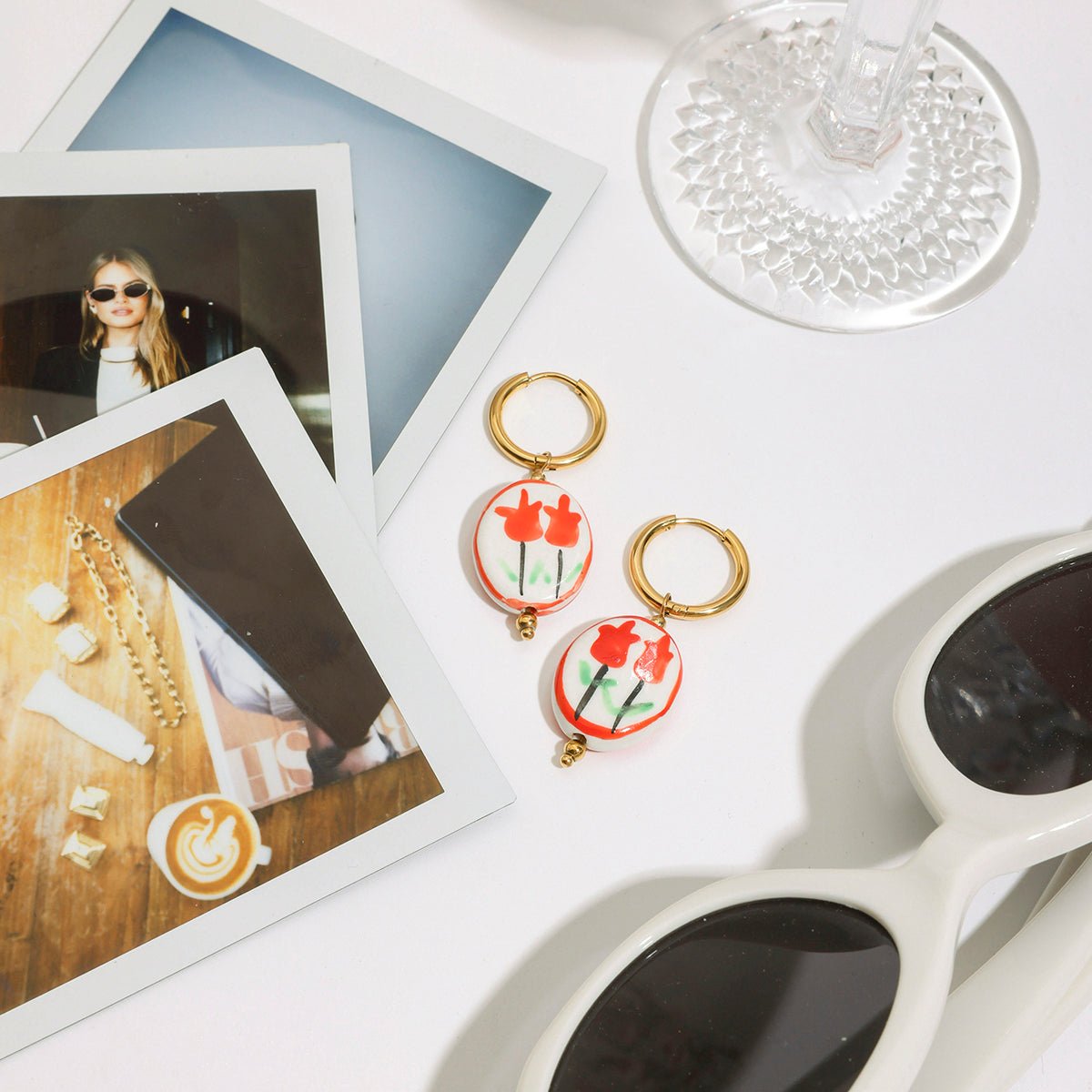 Pair of earrings with red floral design on a white surface with photos and sunglasses.