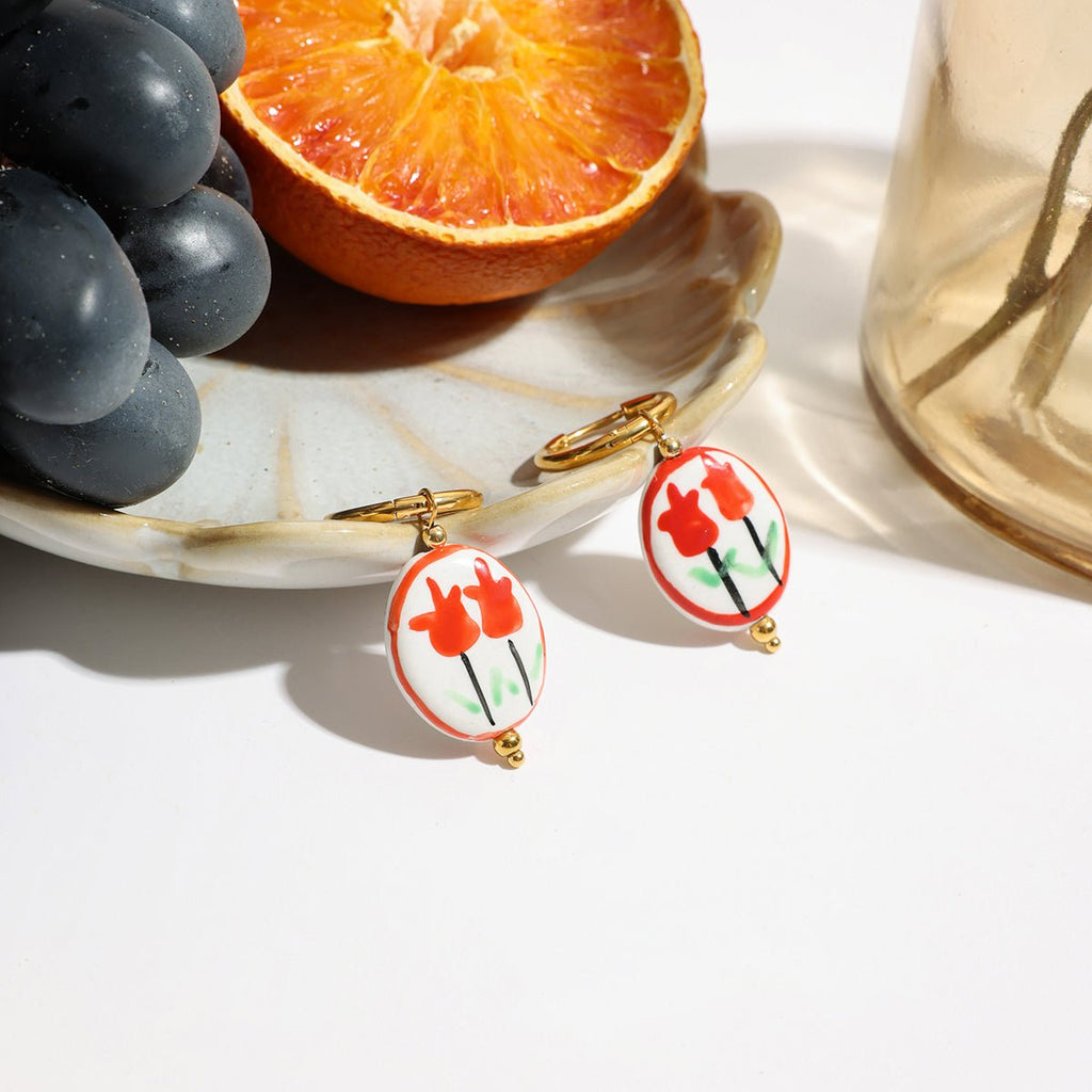 Pair of floral earrings on a white surface with grapes and an orange in the background