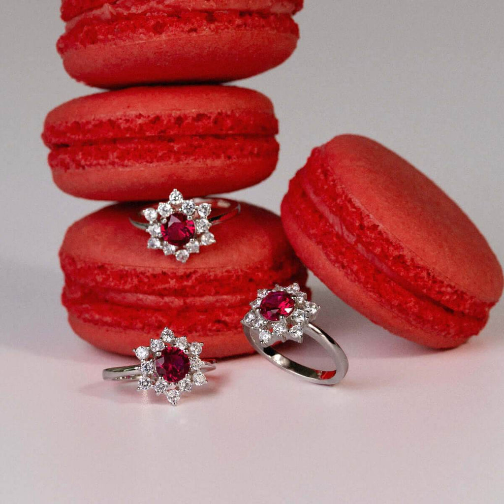 Women’s rhodium-plated silver ring with red ruby-cut cubic zirconia and floral halo design, displayed with red macarons.