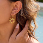 Gold sun-shaped earring worn by a person with a blurred natural background