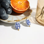 Earrings with blue floral design on a white surface with grapes and an orange.