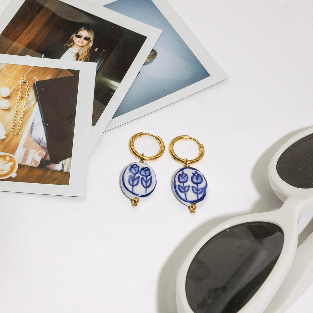 Pair of blue floral earrings with gold accents on a white surface with sunglasses and photos.