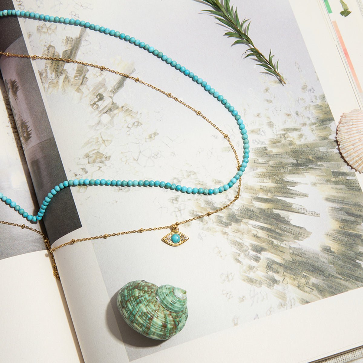 Turquoise beaded necklace with a gold chain and pendant on an open book with a shell and plant leaves.